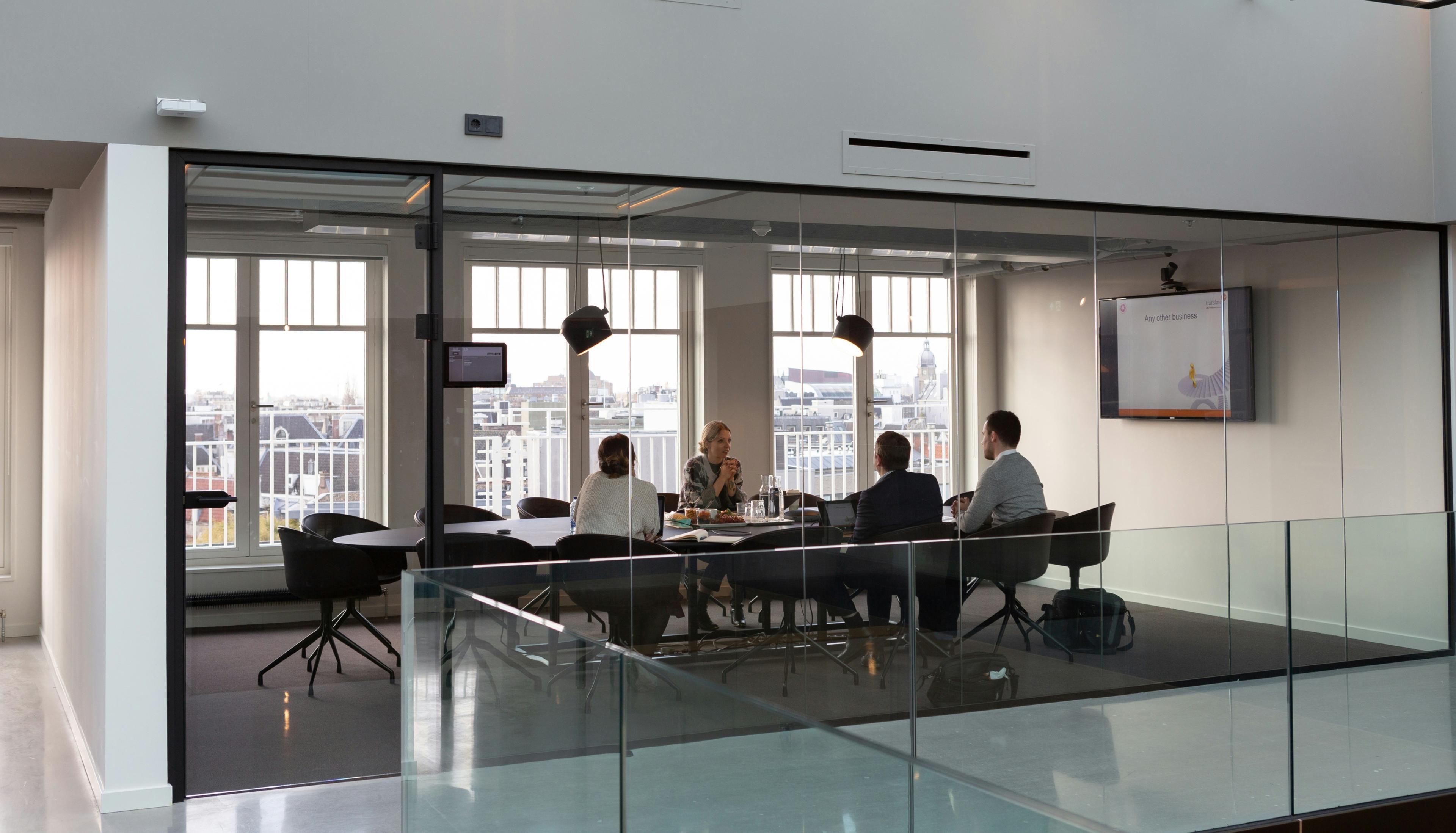 Four people seated around a table in a glass-walled meeting room, engaged in a discussion.