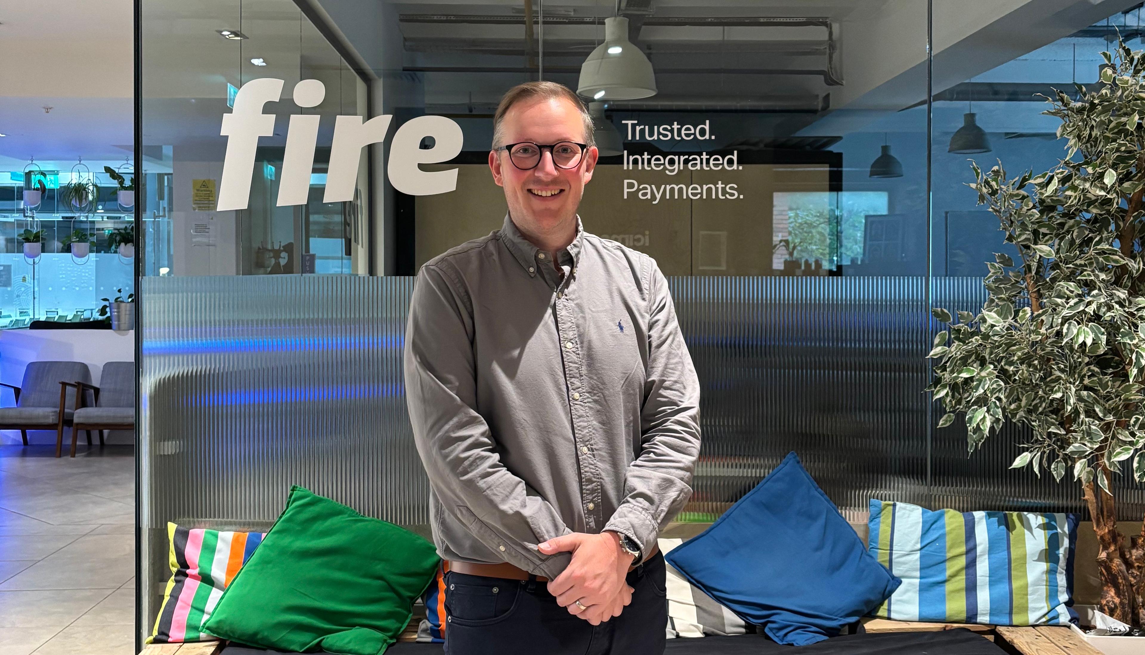 Photo of Patrick Donohoe in front of the glass wall of Fire’s Dublin office. Fire’s logo and the tagline ‘Trusted. Integrated. Payments.’ are visible on the glass wall, and some colourful pillows are on a bench behind Paddy.